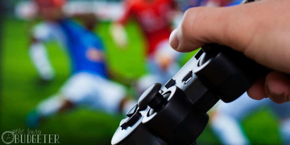 hand holding black gamepad on a front of big screen with soccer game