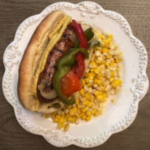 A grilled italian sausage with brown mustard, green pepper, red pepper and onion on a potato roll with a side of roasted corn as one of the easy dinners in the easiest Aldi meal plan, which delivers 14 dinners to your front door for under $150.