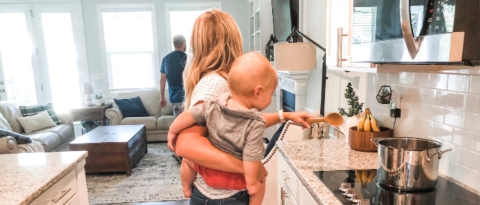 Mom implementing home management routines while she cooks with baby, and talks to husband walking away. 