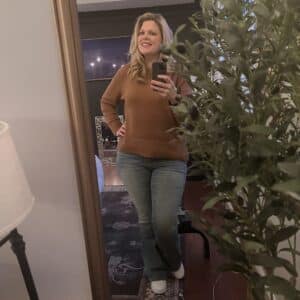 Blonde woman in brown tan turtleneck sweater and grey wash jeans. 