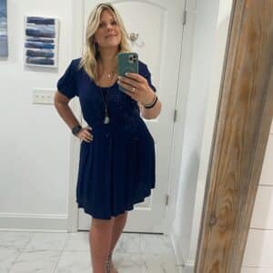 Blonde woman wearingaa navy blue boatneck boho dress with a long pendant. The skirt is mid length. 