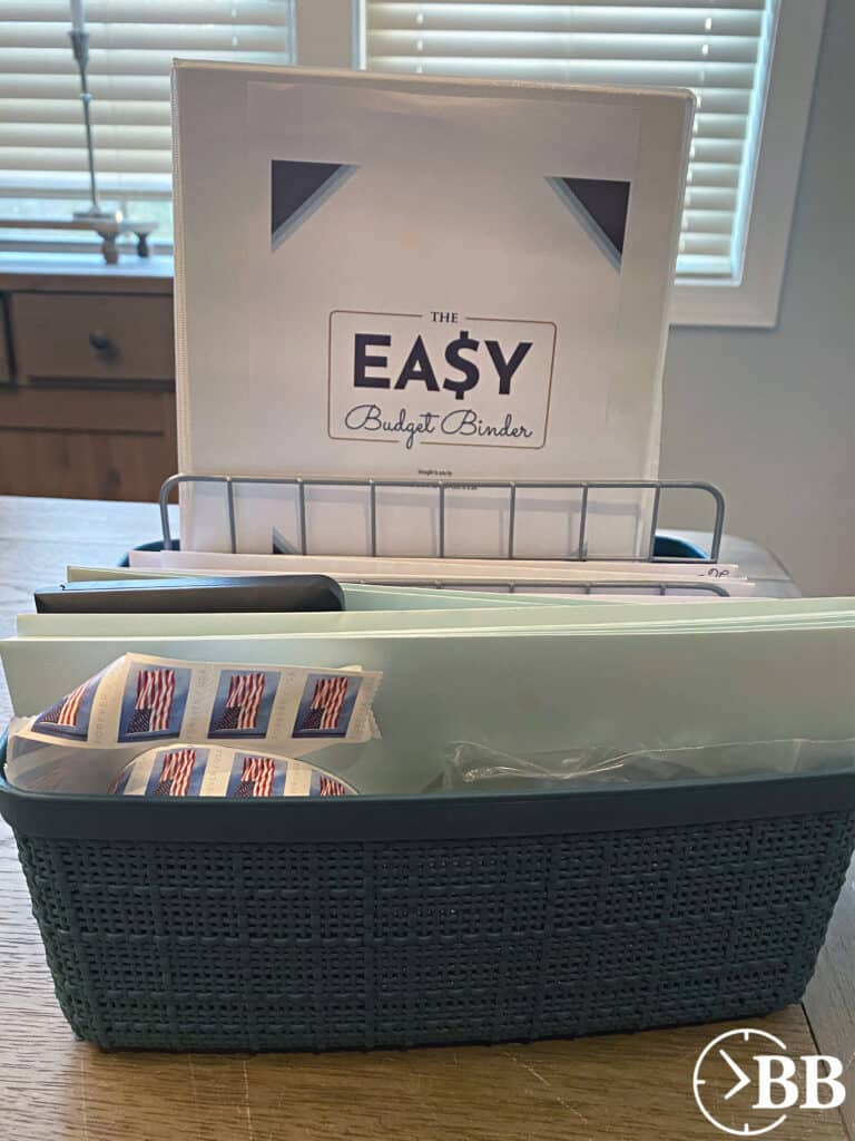 photo of the easy budget binder- a step by step budget system that takes just two minutes a day to maintain. The binder is nestles in a budget box, which has a metal sorter for bills or mail that needs to be dealt with, envelopes, stamps, checks, and pens.