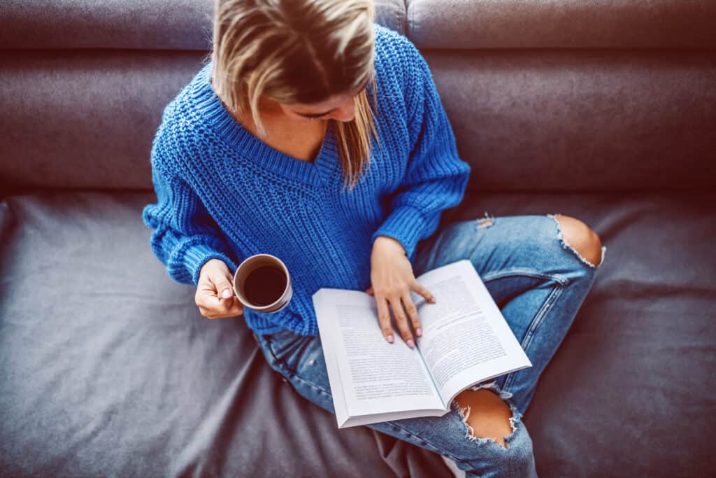 Woman in sweater sitting on sofa in the living room, drinking coffee and reading book.
