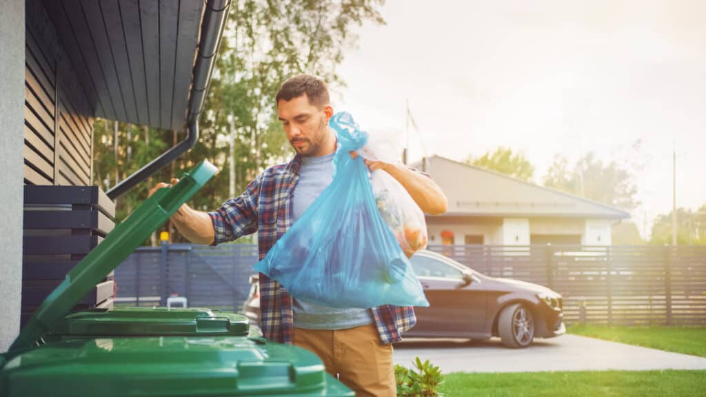 Husband Throwing Away Two Plastic Bags of Trash next to His House. 