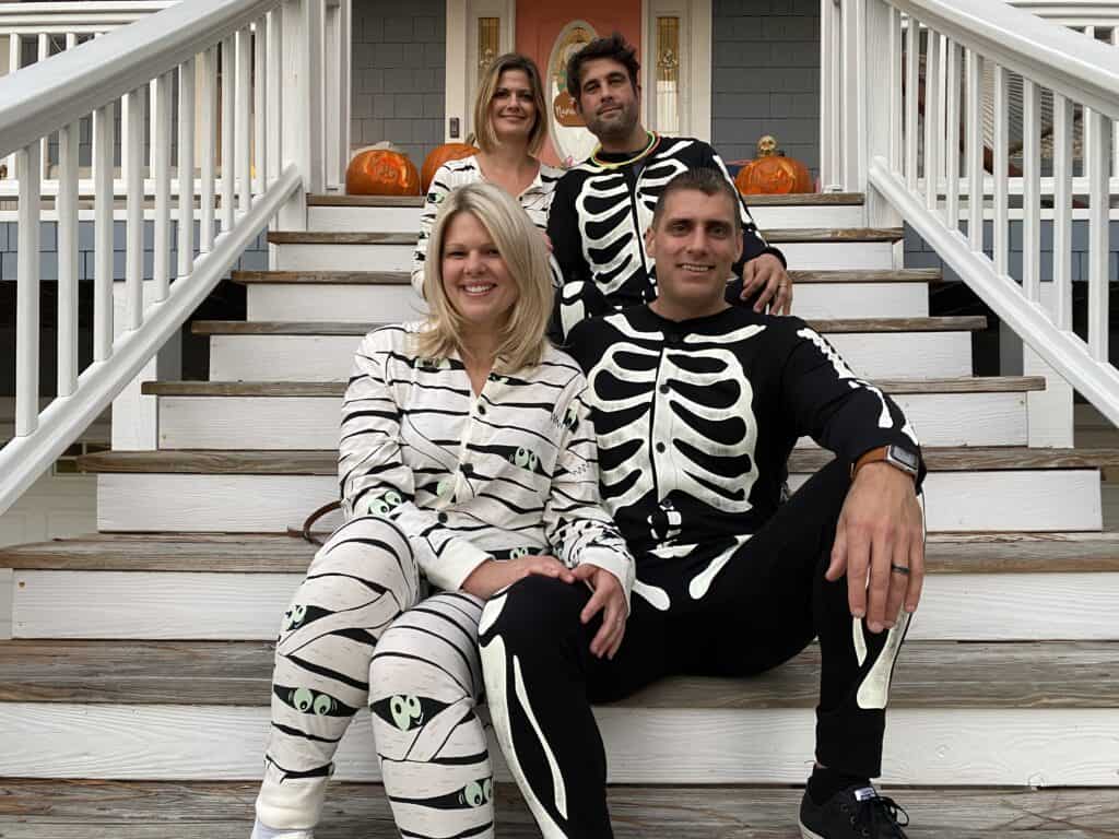 2 couples sitting on the front steps wearing Halloween onesies.