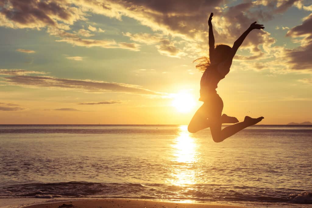 Happy teen girl jumping in the water on the beach at the sunset time