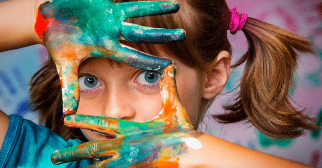 little girl with face framed by paint stained rainbow hands. 