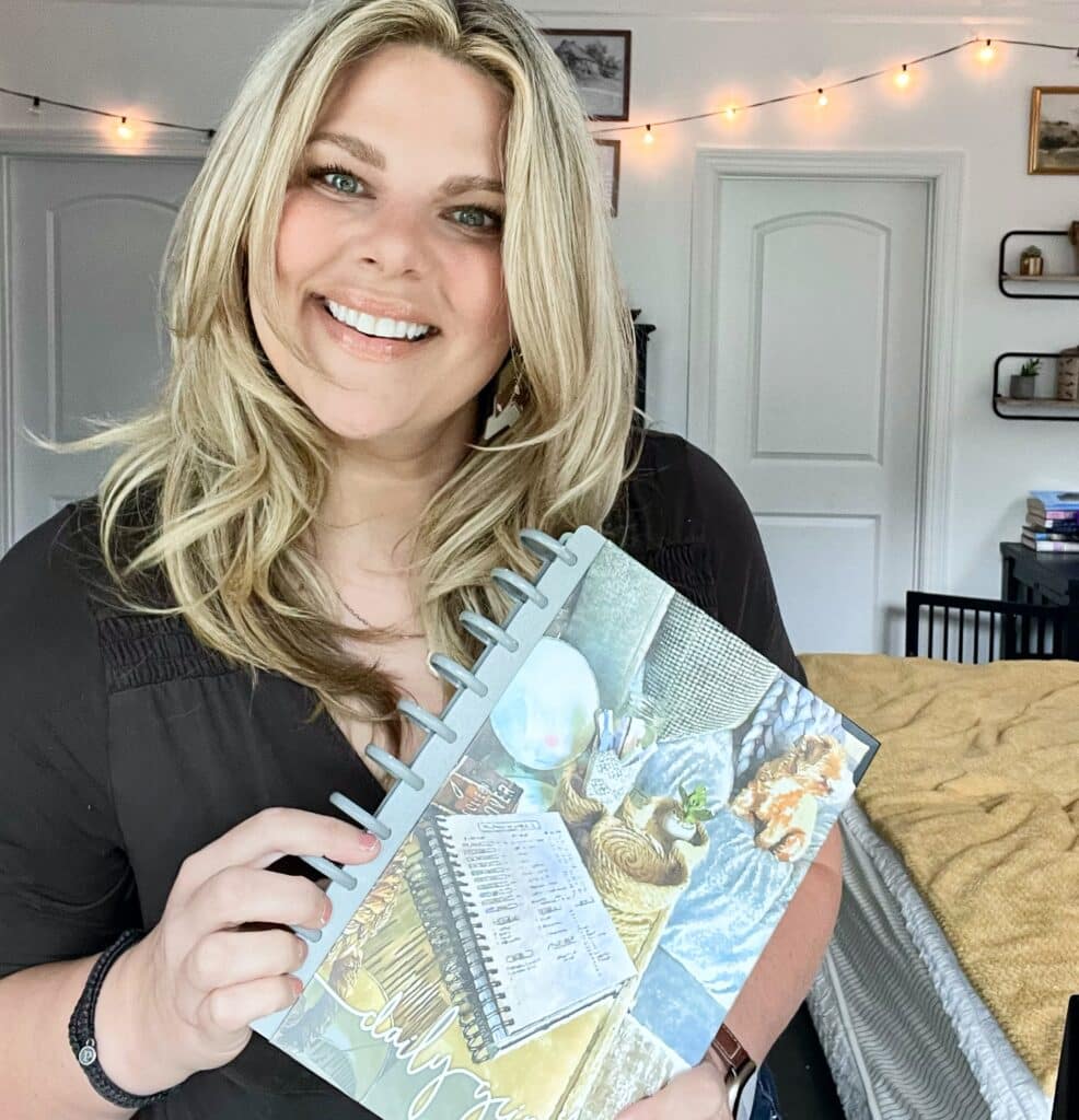 Rosemarie Groner (A blonde woman in a brown shirt) holding the Daily Grind Business Planner up to show off the cover, which is a watercolor painting of the Daily Grinf Life Planner open on a couch with pens and a small dog next to it. 