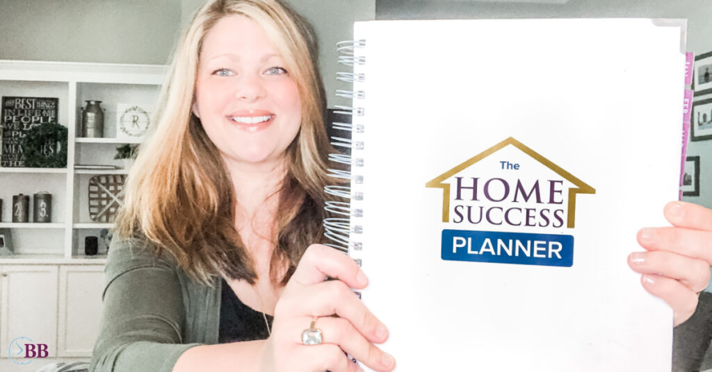 Blonde 40 year old woman smiling holding a planner in front of white built in styled bookshelves. The planner is white and coiled and says "The Home Success Planner" on the front cover of it. 